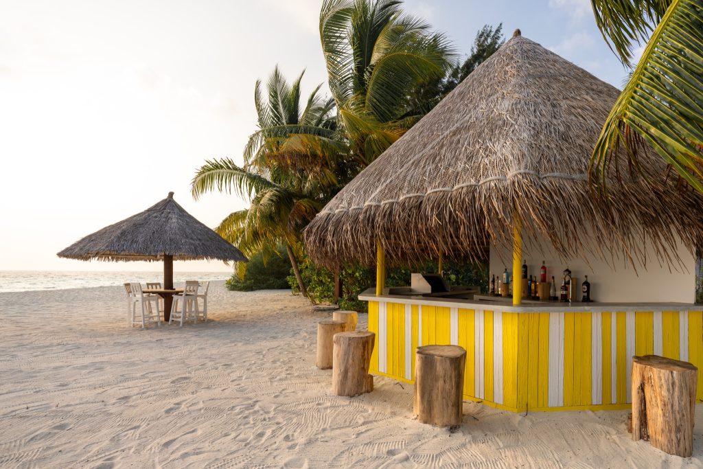 Strandbar mit Strohdach, gelb-weißer Theke und Holzstühlen, daneben runder Tisch unter Strohdach am Strand