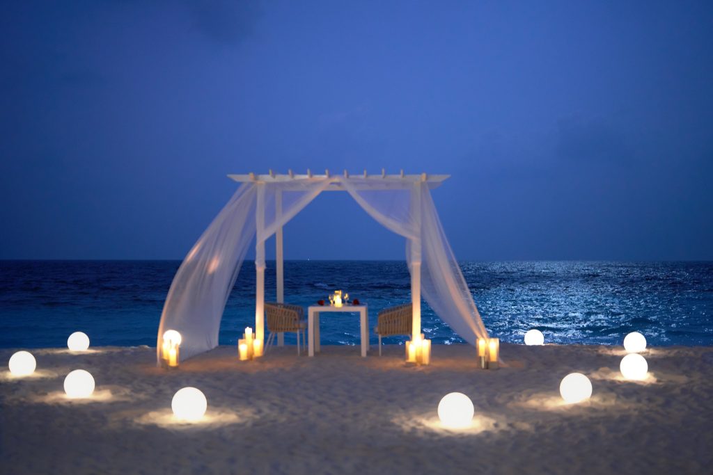 Weiße Holzpergola mit transparenten Vorhängen, Tisch und zwei Stühlen auf hellem Sand, beleuchtet am Strand.