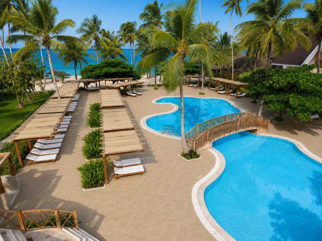 Großer organisch geformter Pool mit klar blauem Wasser, umgeben von Palmen und Holzpavillons in Jaz Amaluna Sansibar