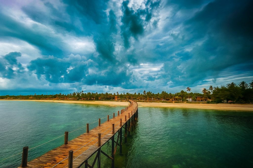 Langer Holzsteg mit Planken und Geländern über grün-blauem Wasser führt zu Sandstrand mit Palmen unter wolkigem Himmel