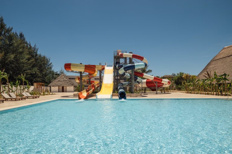 Hotelpool mit roten, gelben, orangen und blauen Wasserrutschen sowie Holzliegen am Rand unter blauem Himmel