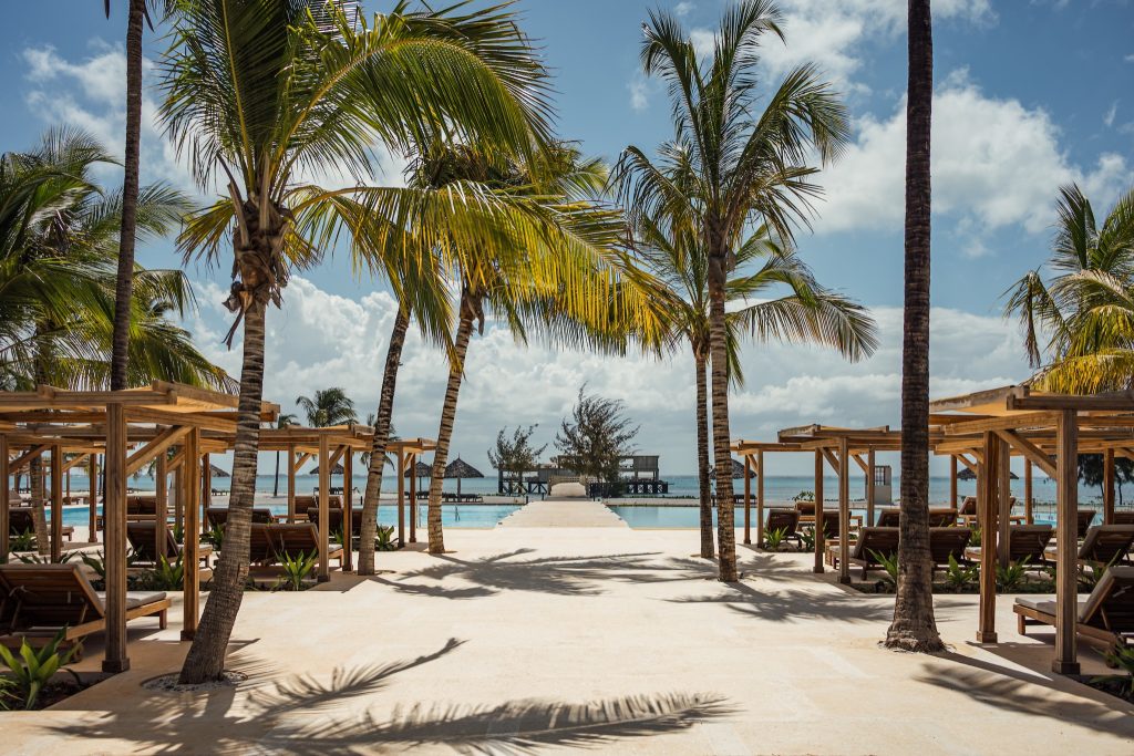Poolbereich mit Holzliegen unter Pavillons, Palmen und Steg zum Ozean im JAZ Elite Aurora Sansibar