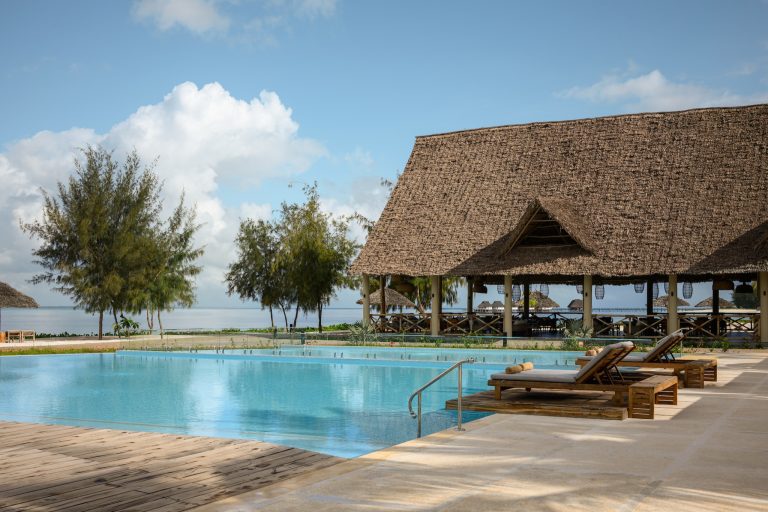 Poolbereich mit klarem Wasser, Holzliegen, Steinfliesen und offenem Pavillon mit Palmblättern am JAZ Elite Aurora Sansibar