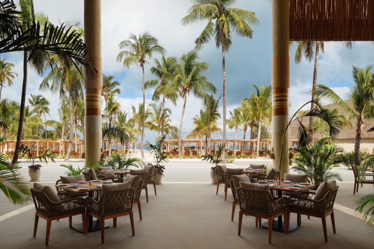 Hotelterrasse des Restaurants Zaafarani mit Holztischen, gepolsterten Sesseln, Säulen und Blick auf tropische Palmen und das.