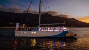 Großer Katamaran mit Passagieren an Deck und innen beleuchtet vor bergiger, bewaldeter Küste bei Abenddämmerung