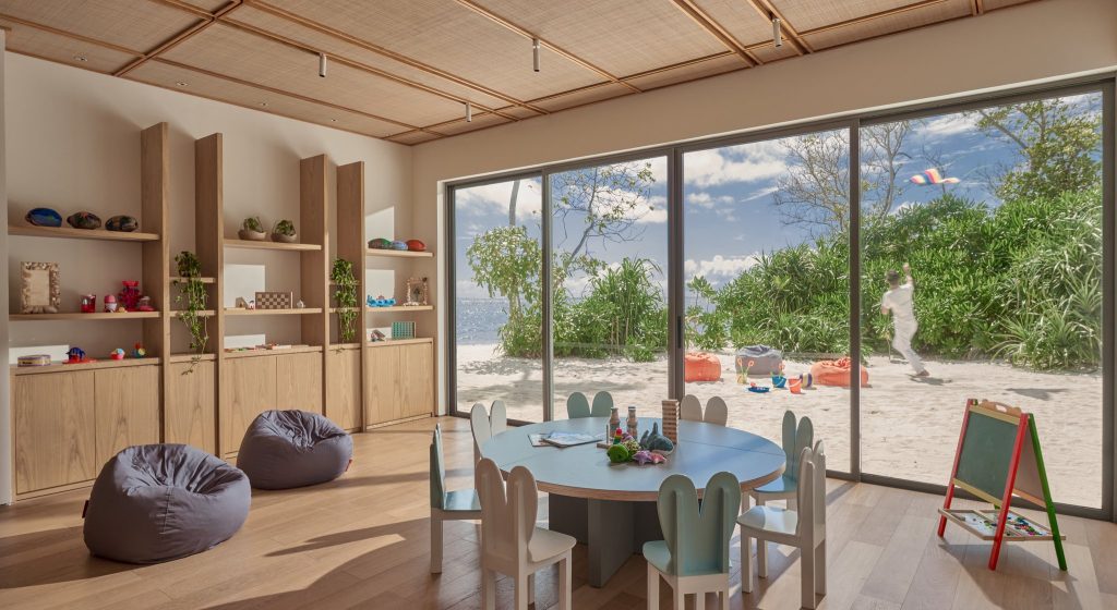 Heller Spielbereich mit rundem Tisch, Holzregalen, Spielzeug und Glasfront mit Blick auf Strand und Meer