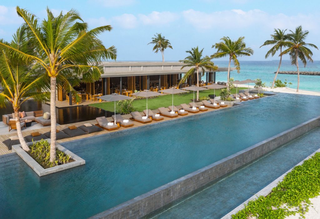 Infinity-Pool mit dunklem Steinrand und gepolsterten Liegen auf Holzterrasse, Palmen und Meer im Hintergrund
