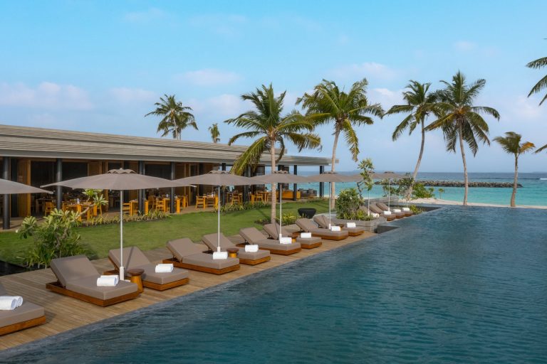 Infinity-Pool mit Holzliegen, beigen Kissen und Sonnenschirmen neben tropischen Pflanzen im Alila Kothaifaru Maldives