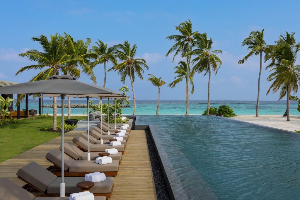 Außenbereich mit Liegestühlen, weißen Handtüchern, grauen Sonnenschirmen, Infinity-Pool und Blick auf das Meer