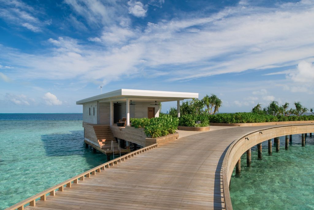 Moderner Hotelbungalow auf Stelzen über türkisblauem Meer mit Holzsteg und Pflanzen umgeben