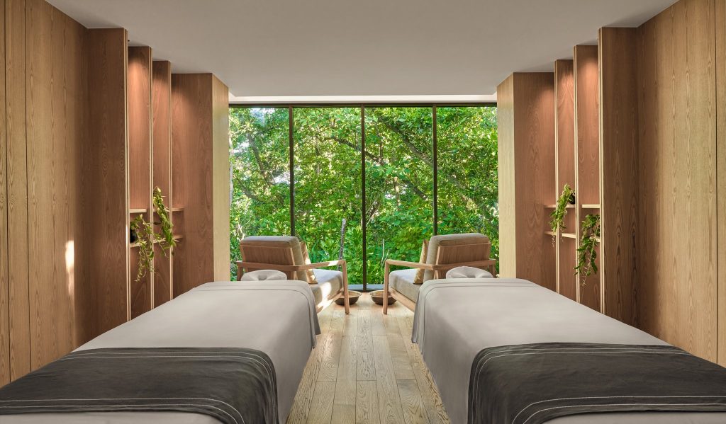 Spa-Raum mit zwei grauen Massageliegen, hellem Holz, Fensterfront und grünem Laub im Hintergrund