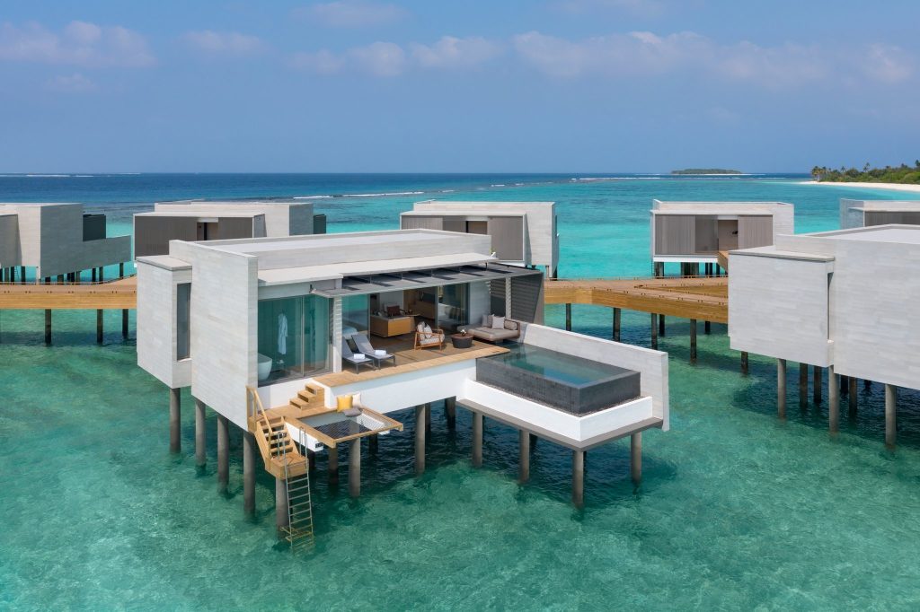 Überwasser-Bungalow mit Glasfront, Holzterrasse, Infinity-Pool und Liegestühlen über türkisfarbenem Wasser