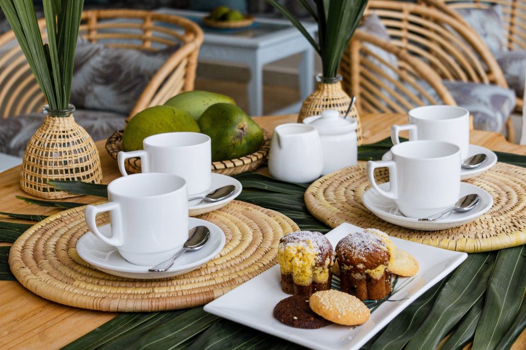 Ein gedeckter Tisch mit weißen Kaffeetassen, Keramikkännchen, Gebäck und Rattanmöbeln in der Bar Chai Lounge
