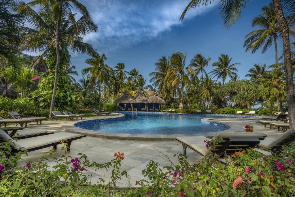 Hotelpool mit Holzliegestühlen, hellen Steinplatten, tropischen Palmen und Strohdachgebäuden im Breezes Beach Club And Spa
