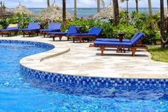 Geschwungener Pool mit blauen Mosaikfliesen, Holzliegen mit blauen Polstern, Palmen und Meer im Hintergrund