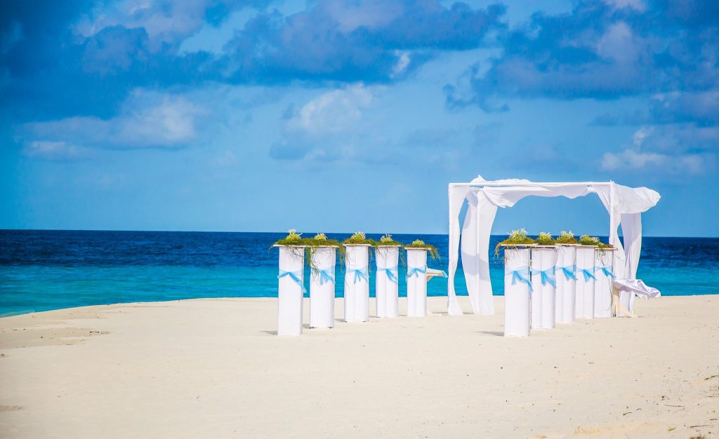 Hochzeitsszene am weißen Sandstrand mit weißen Säulen, blauen Bändern und Hochzeitsbogen vor türkisblauem Meer