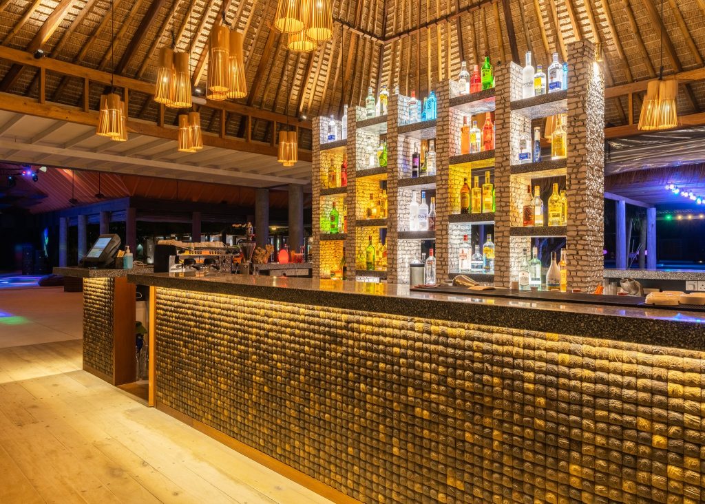 Bar im Furaveri Maldives mit polierter Stein-Theke, kupferfarbenen Lampen und Regal mit bunten Flaschen