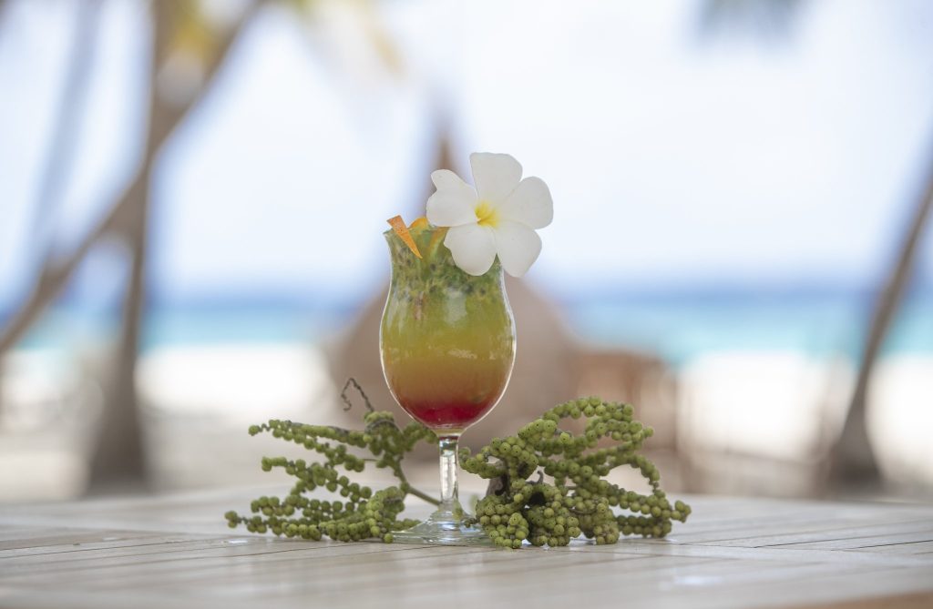 Glas mit farbig geschichtetem tropischem Getränk, dekoriert mit weißer Blüte und oranger Zeste auf Holztisch