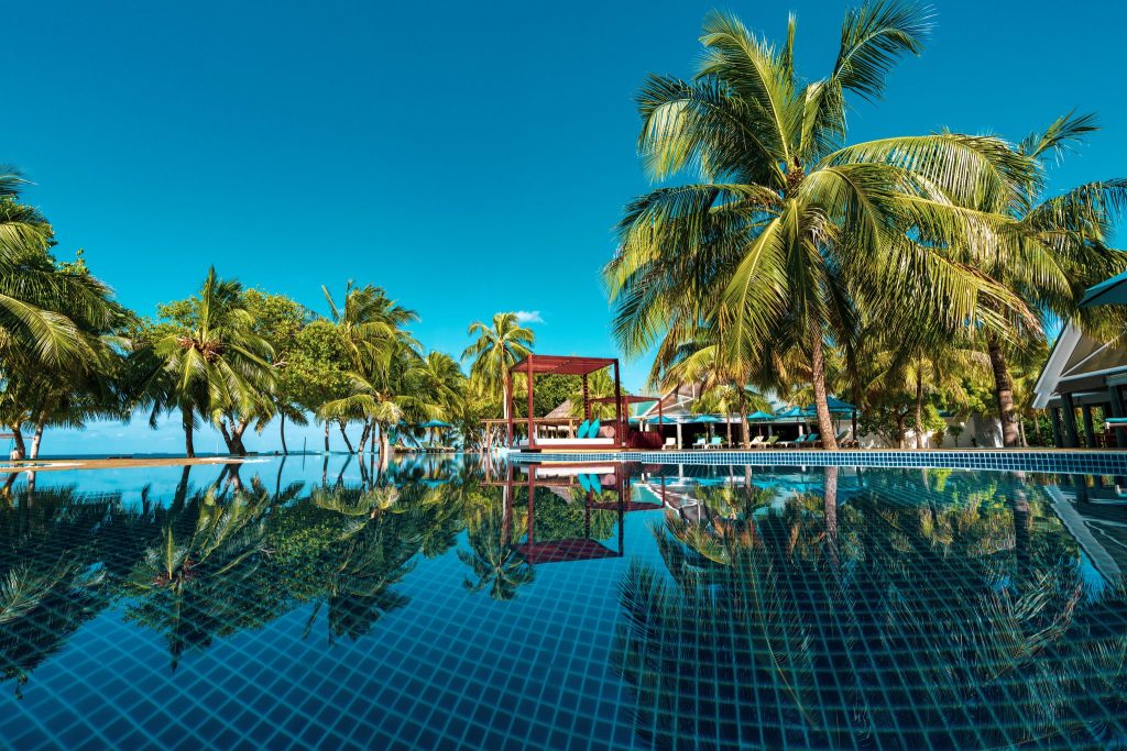 Pool mit klarem Wasser, Holzplattformen, weißen Liegen und Pavillon unter Palmen bei hellem Tageslicht