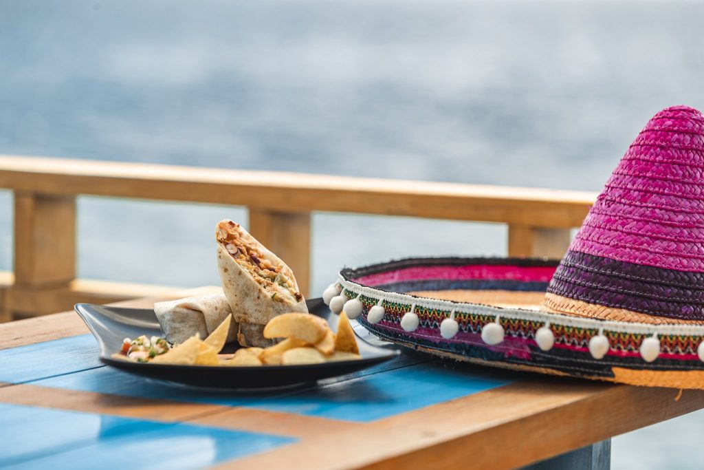 Holztisch mit blauer Platte, Teller mit Wrap, Tortillachips, Salat und farbenfrohem Sombrero, Meer im Hintergrund
