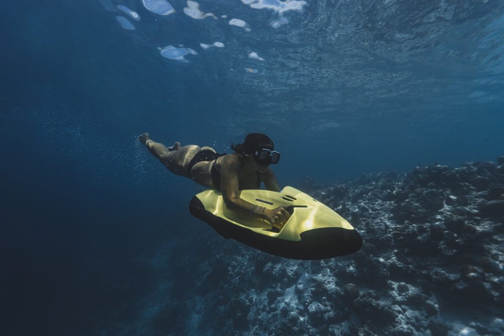 Person fährt unter Wasser auf einem gelben Tauchscooter vor blauem Meeresgrund mit Korallen auf den