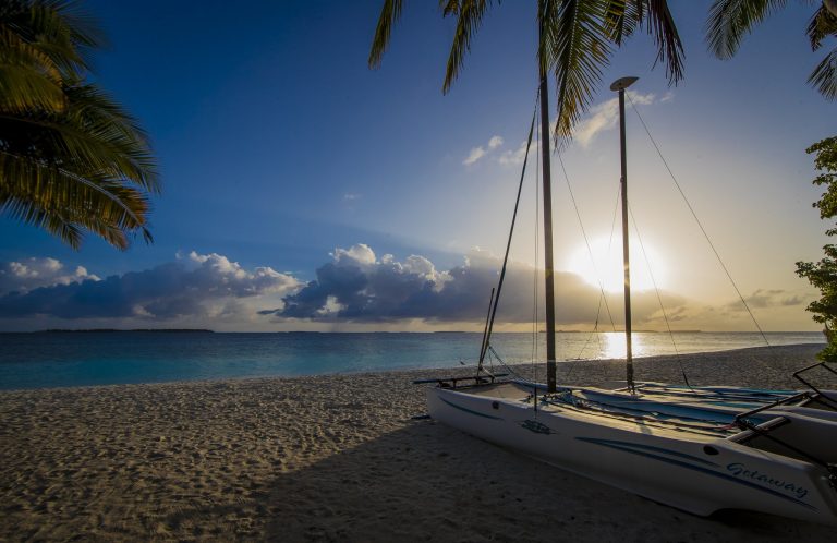 Katamaran aus Glasfaser und Metall auf Sandstrand bei Sonnenuntergang mit Palmen und blauem Himmel auf den