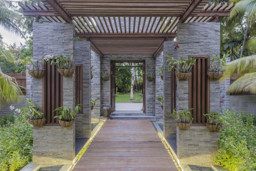 Durchgangsbereich im Furaveri Maldives Wellness Spa mit grauen Steinsäulen, hölzerner Pergola und grünen Pflanzen