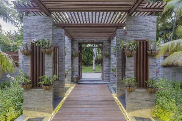 Durchgangsbereich im Furaveri Maldives Wellness Spa mit grauen Steinsäulen, hölzerner Pergola und grünen Pflanzen
