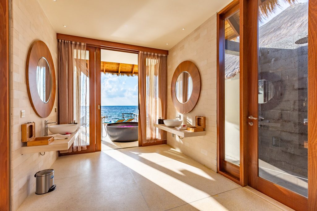 Badezimmer mit zwei runden Spiegeln, modernen Waschbecken auf Holz, sandfarbenem Boden und Terrasse mit Badewanne.
