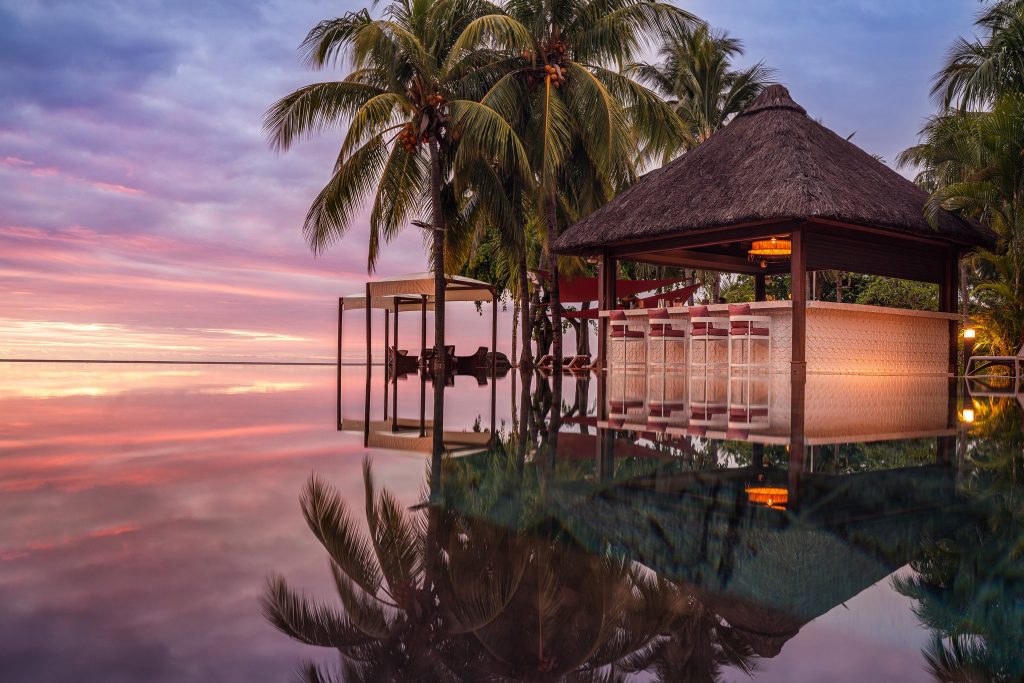 Tropische Bar Hibiscus mit Strohdach am Infinity-Pool, Palmen und Sonnenuntergangshimmel bei Hilton Mauritius Resort & Spa