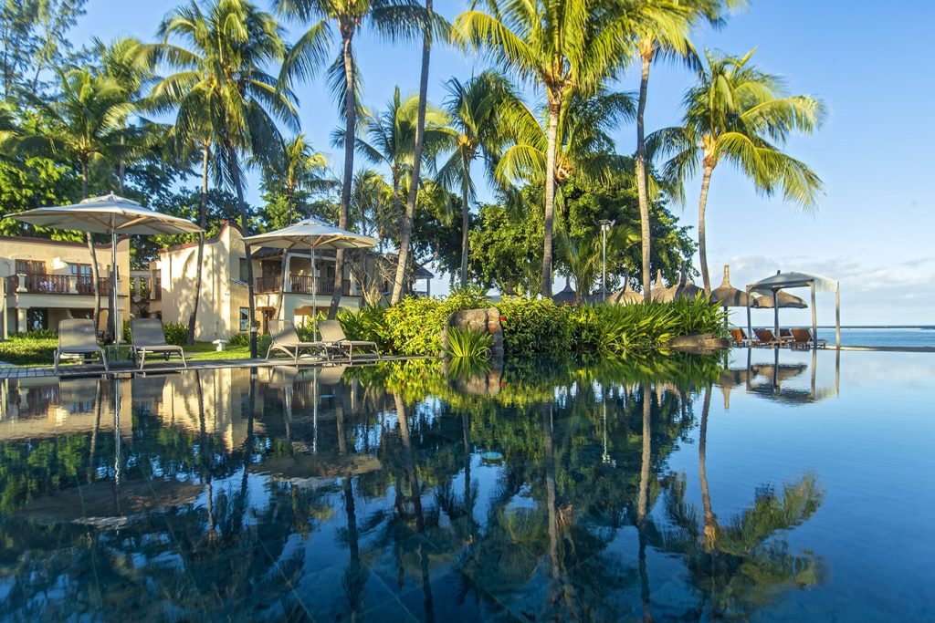 Hotelpool im Hilton Mauritius Resort & Spa mit Liegestühlen, weißen Sonnenschirmen und sandfarbenem Hotelgebäude