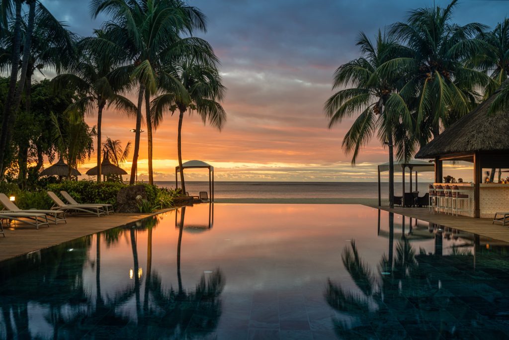 Infinity-Pool mit Sonnenliegen auf Holzdeck, Palmen und Strohpavillon bei Sonnenuntergang im Hilton Mauritius Resort & Spa