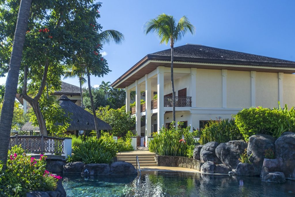 Pool mit klarem Wasser, natürlichen Felssteinen und tropischen Pflanzen vor cremefarbenem Hotelgebäude