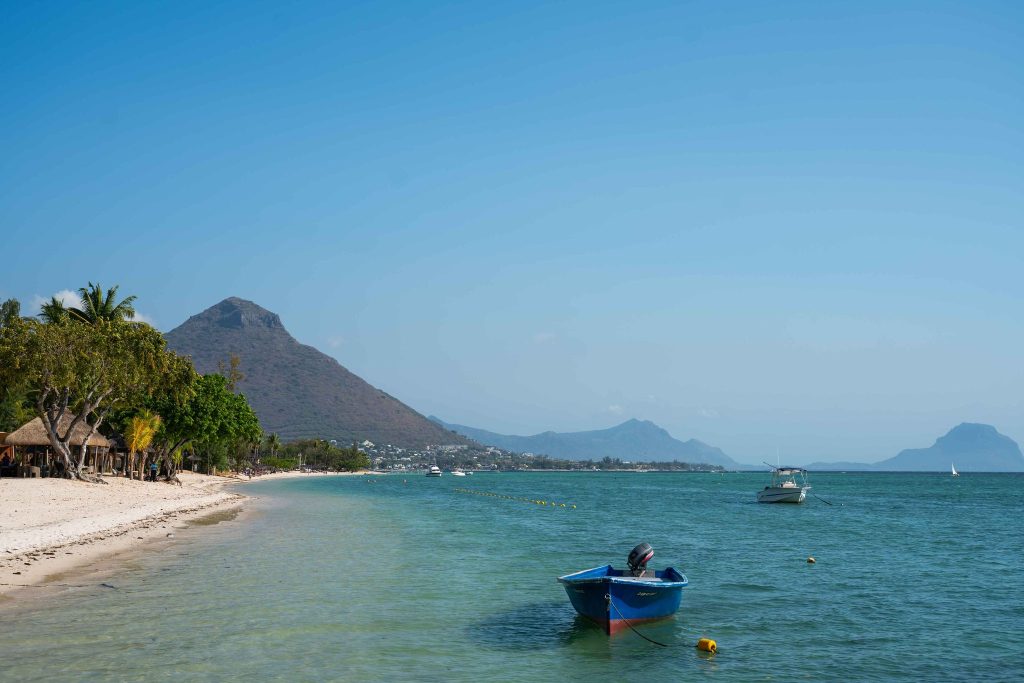 Strand mit hellem Sand, Palmen, blauem Boot im türkisfarbenen Wasser und Bergen im Hintergrund