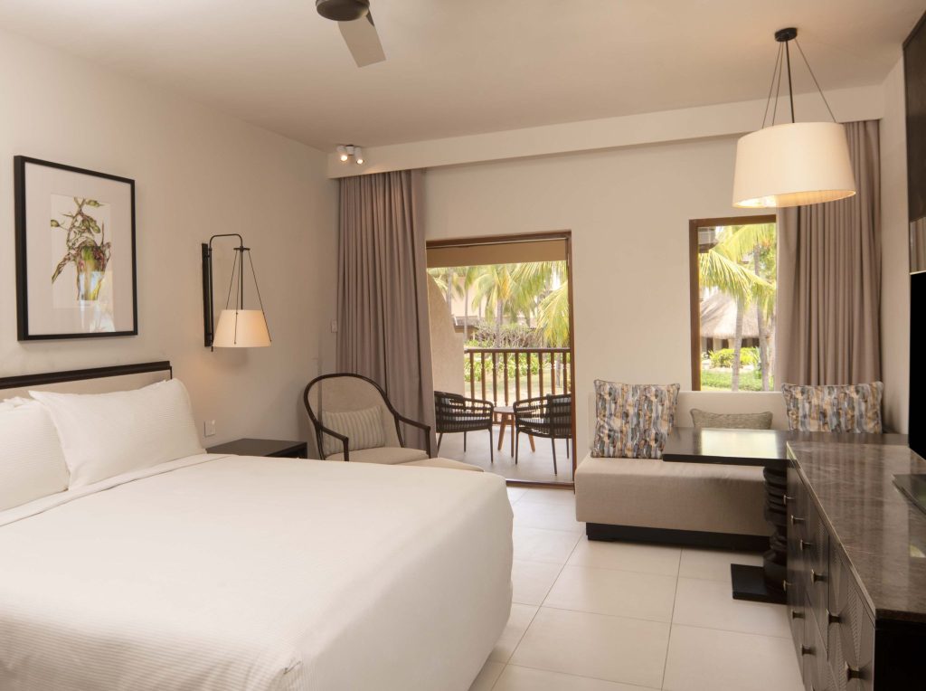 Grand Deluxe Room im Hilton Mauritius mit großem Bett, Sofa, Esstisch und Blick auf Balkon mit Palmen
