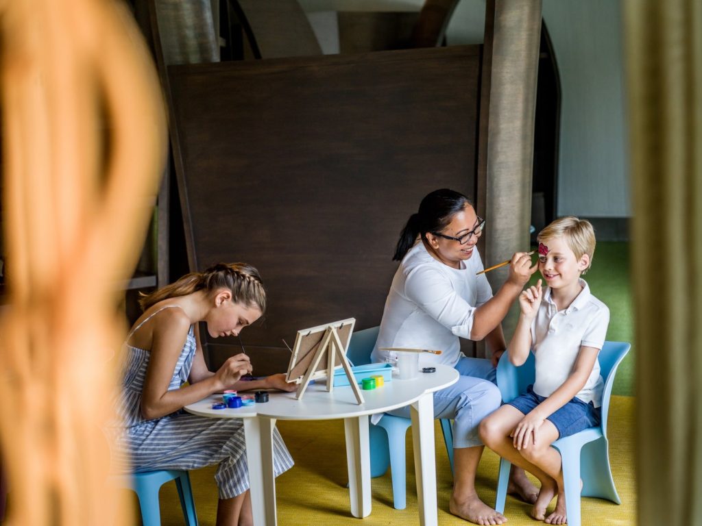 Innenraum mit zwei Kindern und einem Erwachsenen, Gesichtsbemalung und Malen auf Tisch im Resort