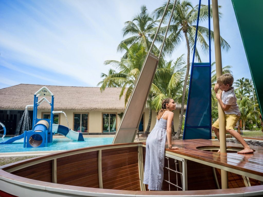 Holzspielgerät in Form eines Schiffsdecks mit spielenden Kindern vor Wasserpark mit blauen Rutschen und Strohdachgebäude