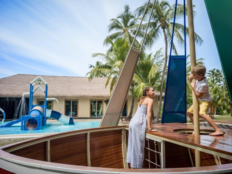 Holzspielgerät in Form eines Schiffsdecks mit spielenden Kindern vor Wasserpark mit blauen Rutschen und Strohdachgebäude