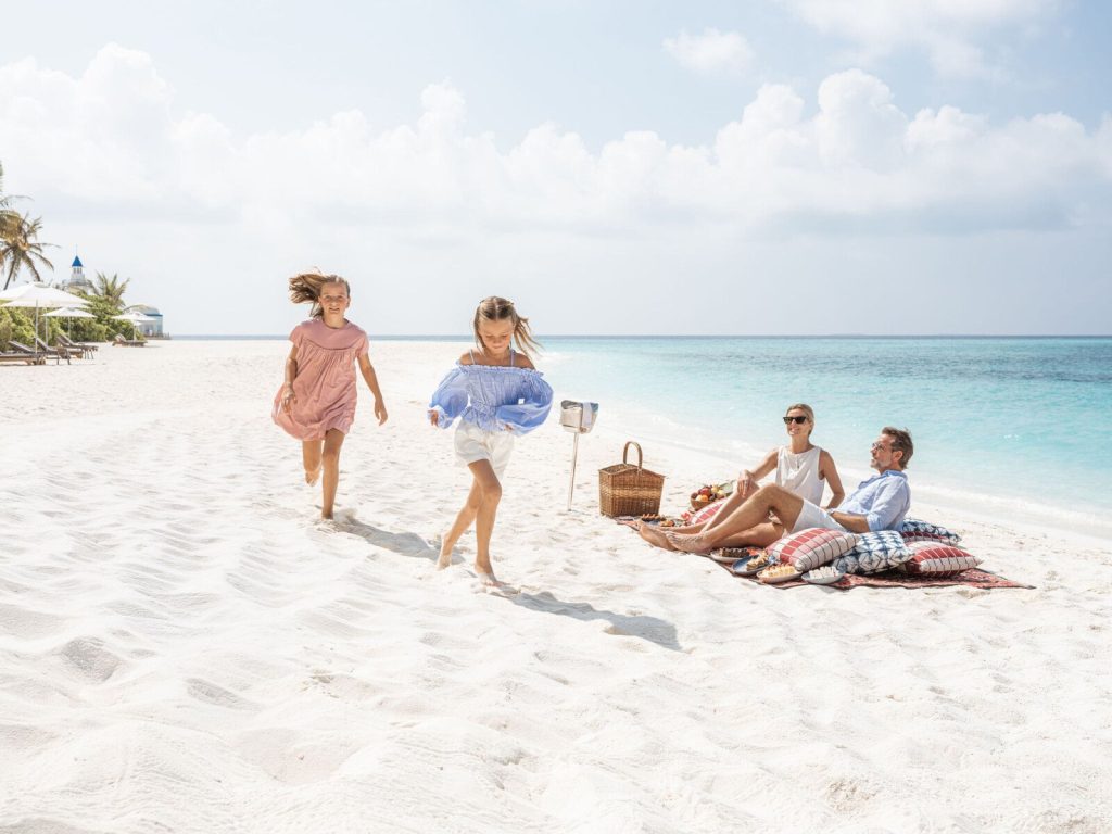 Zwei Kinder laufen barfuß über weißen Sandstrand, zwei Erwachsene sitzen auf bunter Decke mit Picknickkorb.