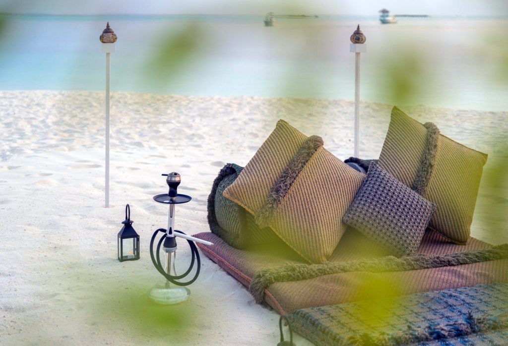 Strand mit gestreiften Kissen in Beige- und Brauntönen, metallische Wasserpfeife und Laterne vor türkisfarbenem Meer