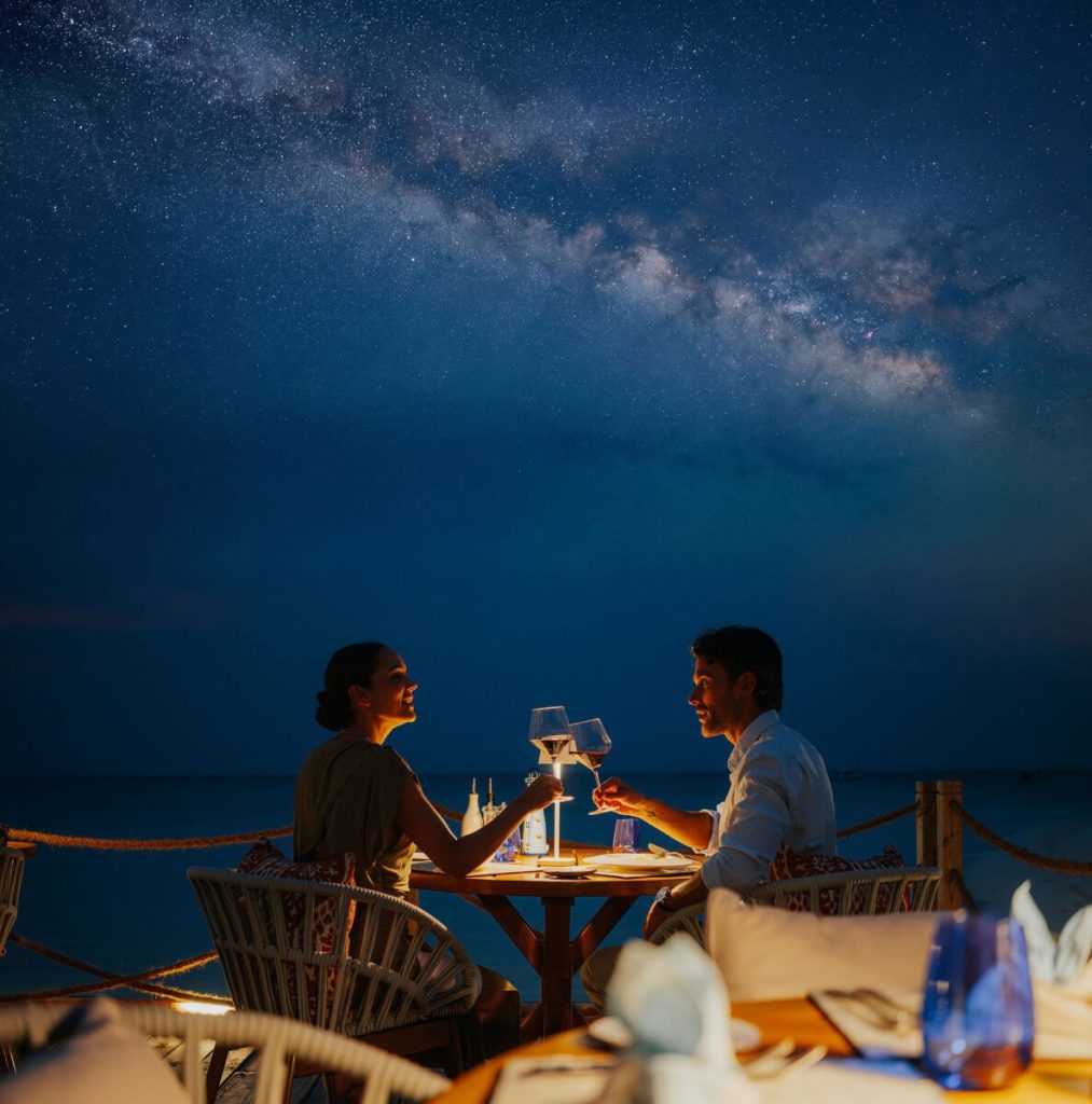 Zwei Personen sitzen an einem Holz-Tisch mit Weinflaschen unter Sternenhimmel im InterContinental Maldives Maamunagau Resort