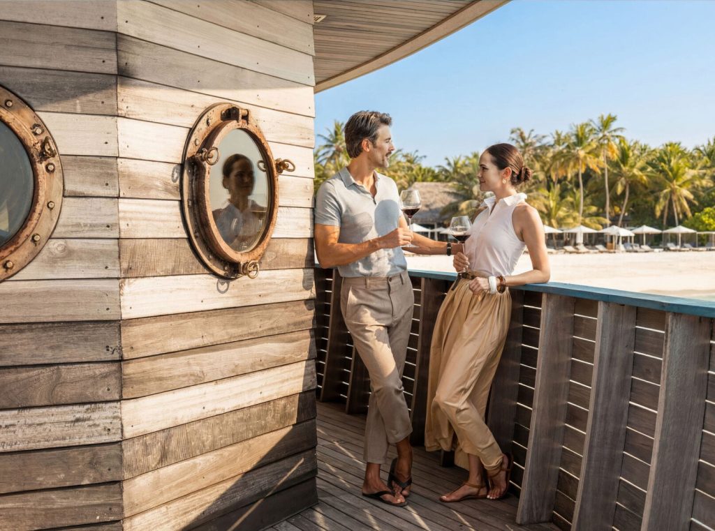 Balkon aus hellem Holz mit Bullaugenfenstern und zwei Personen beim Wein auf den