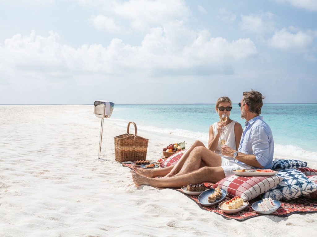 Zwei Personen sitzen auf buntem Teppich mit Kissen und Picknickkorb am weißen Sandstrand