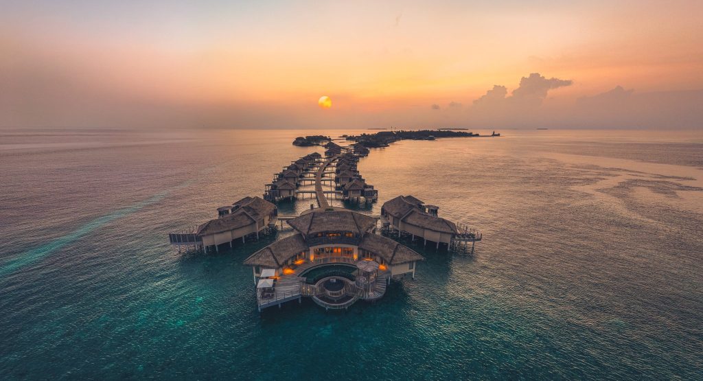 Luftaufnahme von über Wasser gebauten Bungalows mit Strohdächern und Stegen im InterContinental Maldives Maamunagau Resort