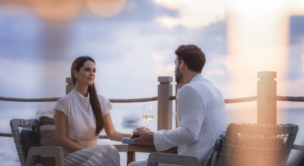 Zwei Personen sitzen an Holztisch in der sunset-bar mit Geländer aus Seilen und Blick auf das Meer