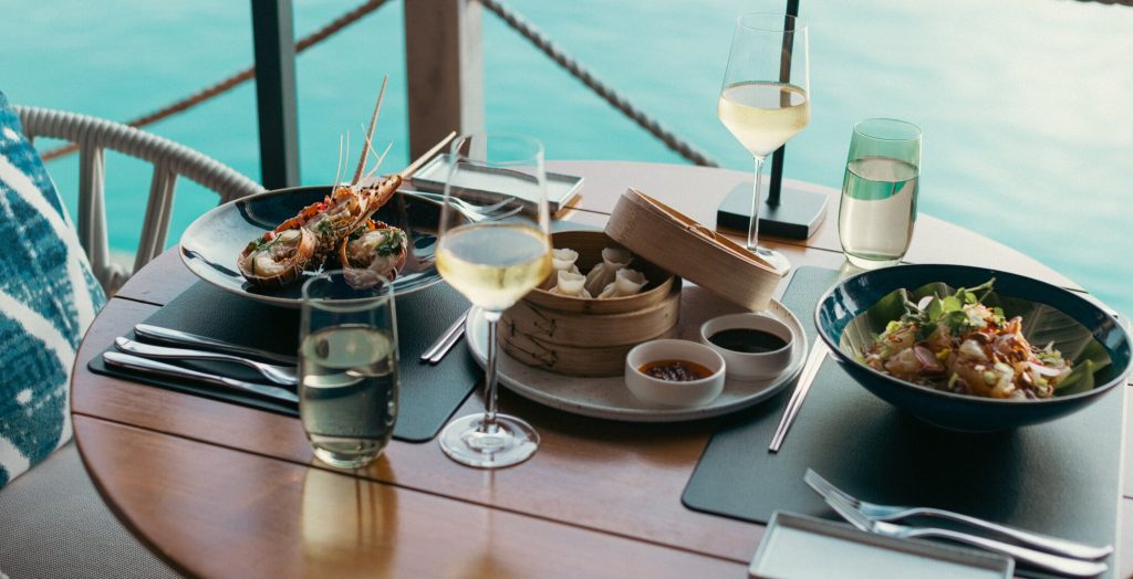 Runder Holztisch mit Meeresfrüchten, Dumplings, Salat, zwei Gläsern Weißwein, türkisem Wasser im Hintergrund