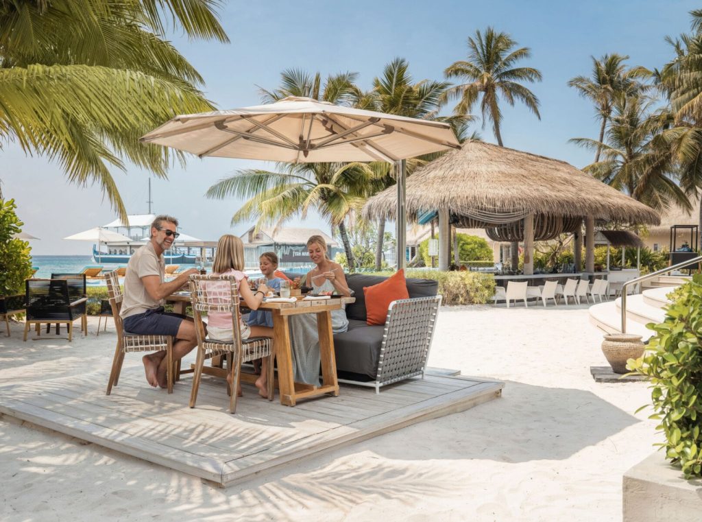 Outdoor-Dining-Bereich im Restaurant-Café Umi mit Holztisch, Palmen, strohgedecktem Gebäude und Ozean im Hintergrund