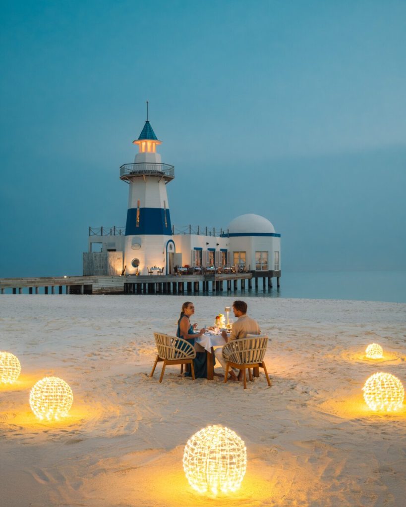 Gedeckter Esstisch am Strand mit zwei Stühlen und blau-weißem Leuchtturm im Hintergrund bei Dämmerung