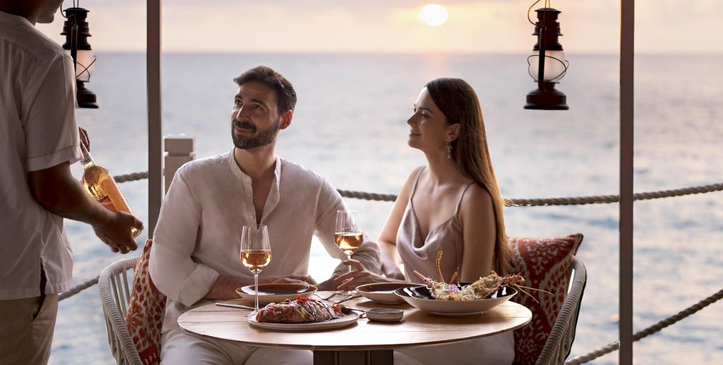 Zwei Personen sitzen an rundem Holztisch im Restaurant Fish Market mit Speisen, Weingläsern und Laternen am Meer