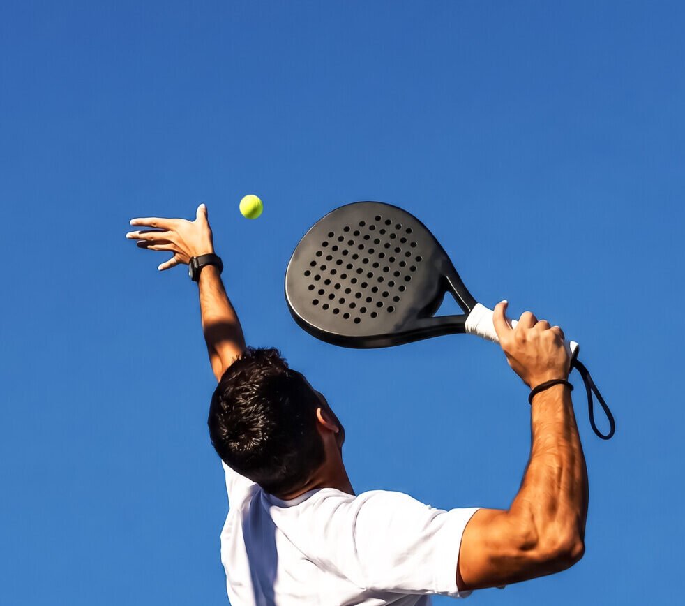 Person mit weißem Oberteil beim Padel-Tennis Schlag mit schwarzem Schläger und gelbem Ball vor blauem Himmel